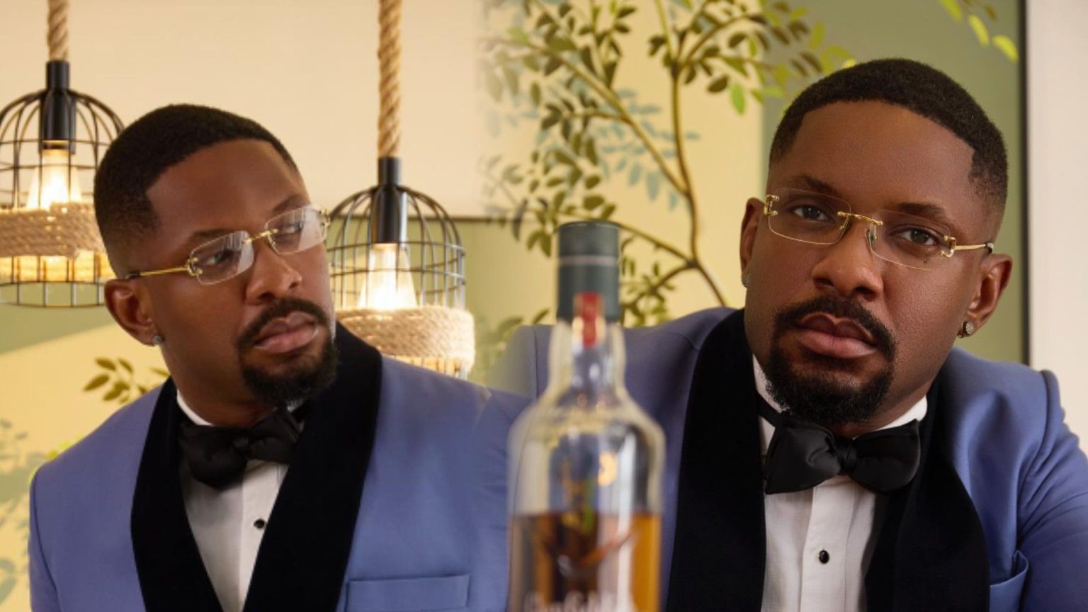 Cross Okonkwo, popularly known as BBNaija Cross, poses in a sharp blue tuxedo with black lapels, wearing gold-rimmed glasses and a bow tie. The background shows modern hanging lamps and leafy wall art, creating a refined indoor setting.