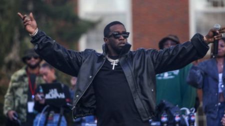 Diddy performs on stage in a black leather jacket and sunglasses, raising both arms before a crowd.