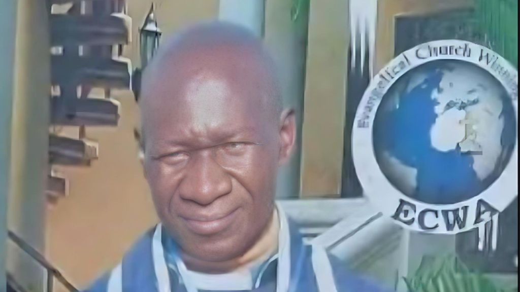 Portrait of Rev Anthony Lamba wearing a blue clerical robe, standing in front of an Evangelical Church Winning All (ECWA) emblem.