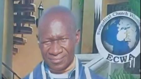 Portrait of Rev Anthony Lamba wearing a blue clerical robe, standing in front of an Evangelical Church Winning All (ECWA) emblem.