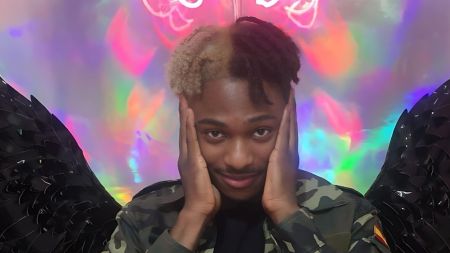 BBNaija housemate Faith poses with his hands on his face, wearing a camouflage jacket. He has half-dyed hair, with one side blonde and the other black, and black feathered wings in the background against a colourful neon backdrop.