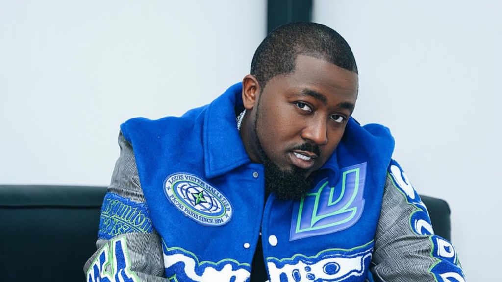 Nigerian rapper Ice Prince poses in a stylish blue Louis Vuitton varsity jacket while seated, looking directly at the camera.