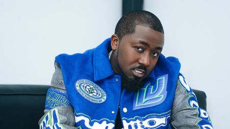 Nigerian rapper Ice Prince poses in a stylish blue Louis Vuitton varsity jacket while seated, looking directly at the camera.