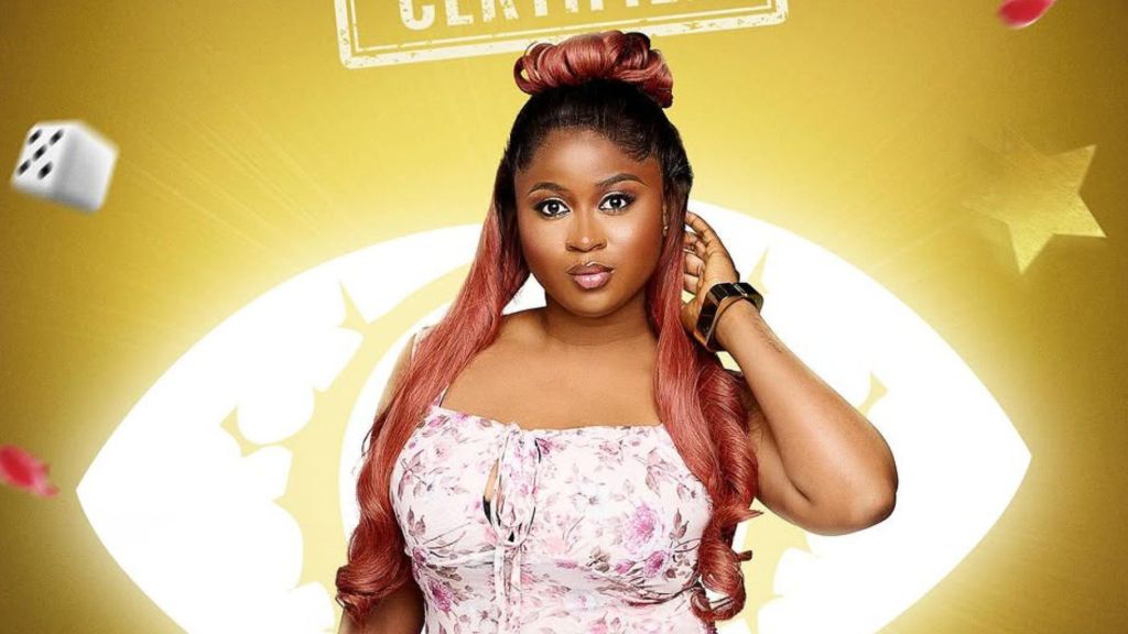 BBNaija housemate Imisi poses confidently against a golden backdrop with the show’s eye logo. She wears a floral pink dress, styled with long wavy reddish hair and a bold wristwatch, while gently holding her hair with one hand.