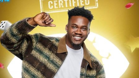 BBNaija housemate Jason poses against the golden certified background, smiling brightly and pointing at himself while wearing a striped jacket over a white shirt.