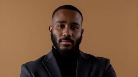 BBNaija star Kiddwaya poses in a black suit against a plain brown background.