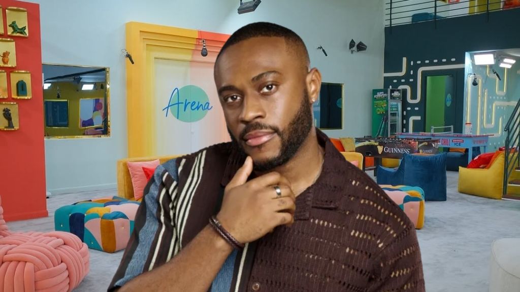 BBNaija Season 10 housemate Kola poses confidently inside the Big Brother Naija house lounge, wearing a brown patterned shirt with his hand on his chin.