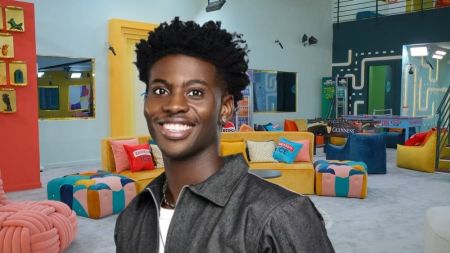 BBNaija Season 10 housemate Koyin smiles brightly while posing inside the Big Brother Naija house lounge, wearing a dark jacket with a white shirt underneath.