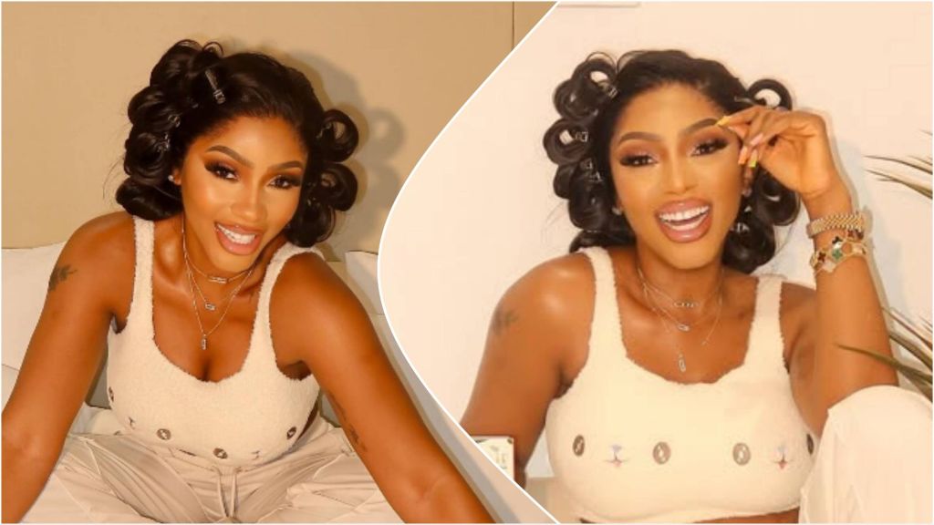 BBNaija star Mercy Eke smiling brightly while seated on a bed, wearing a cream-coloured sleeveless top and matching trousers, with her hair styled in loose curls.