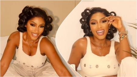 BBNaija star Mercy Eke smiling brightly while seated on a bed, wearing a cream-coloured sleeveless top and matching trousers, with her hair styled in loose curls.