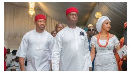 Michael Nwoko, Senator Ned Nwoko, and Regina Daniels walk together at an indoor event dressed in traditional white Igbo attire, with Regina wearing coral beads and a white head wrap.