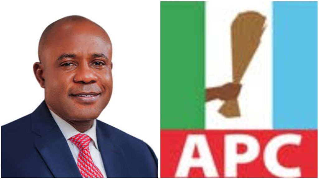 Enugu State Governor Peter Mbah is pictured wearing a dark blue suit with a red patterned tie, smiling confidently beside the official logo of Nigeria’s ruling party, the All Progressives Congress (APC), which features a hand holding a broom over green, white, and blue stripes.