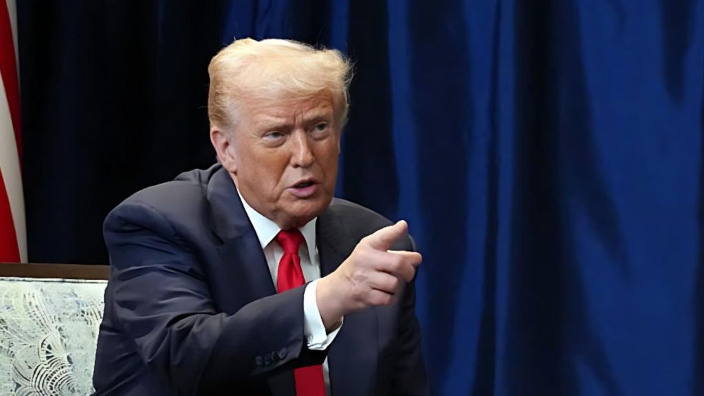 Former US President Donald Trump sits in a suit and red tie, pointing forward while speaking, with a dark blue curtain behind him.