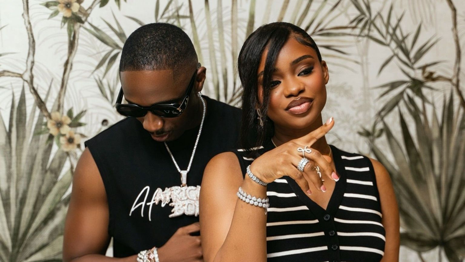 Priscilla Ojo stands in front smiling and flashing her jewellery while Juma Jux, wearing dark sunglasses and a sleeveless black top, stands slightly behind her with his head bowed; leafy wallpaper is in the background.