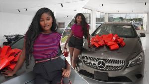 Raye poses proudly beside her new grey Mercedes-Benz sedan decorated with a large red bow, inside a bright showroom with white walls and glass windows.