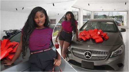 Raye poses proudly beside her new grey Mercedes-Benz sedan decorated with a large red bow, inside a bright showroom with white walls and glass windows.