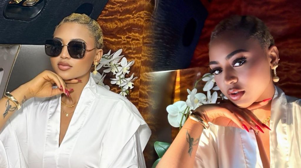 Regina Daniels poses elegantly in a luxury setting wearing a white silk robe, gold accessories, and sunglasses, with white orchids in the background.