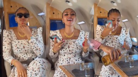 Regina Daniels smiles and pours a drink while seated inside a private jet, wearing a white patterned dress and gold jewellery.