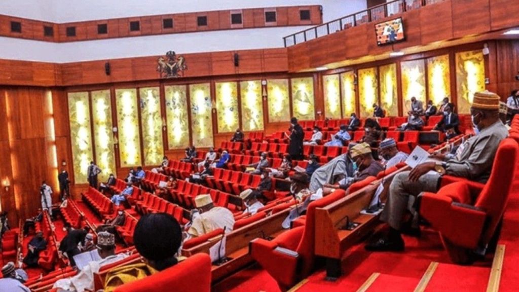 Interior view of the Nigerian Senate chamber in Abuja showing lawmakers seated on red chairs during a plenary session, with some standing and engaging in discussions.