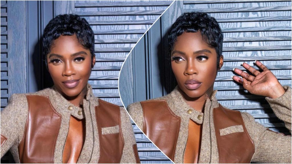 Nigerian singer Tiwa Savage poses confidently in a brown-leather and tweed outfit, showcasing her short wavy hairstyle against a wooden backdrop.