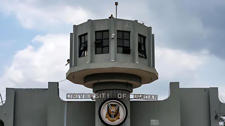 The main gate of the University of Ibadan, featuring its iconic white and grey tower structure with the university’s emblem at the centre, set against a cloudy blue sky.