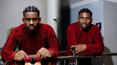 BBNaija housemate Victory Okokon poses in a red textured sweater, styled with neatly braided cornrows and holding a pair of red-tinted sunglasses while gazing confidently at the camera.