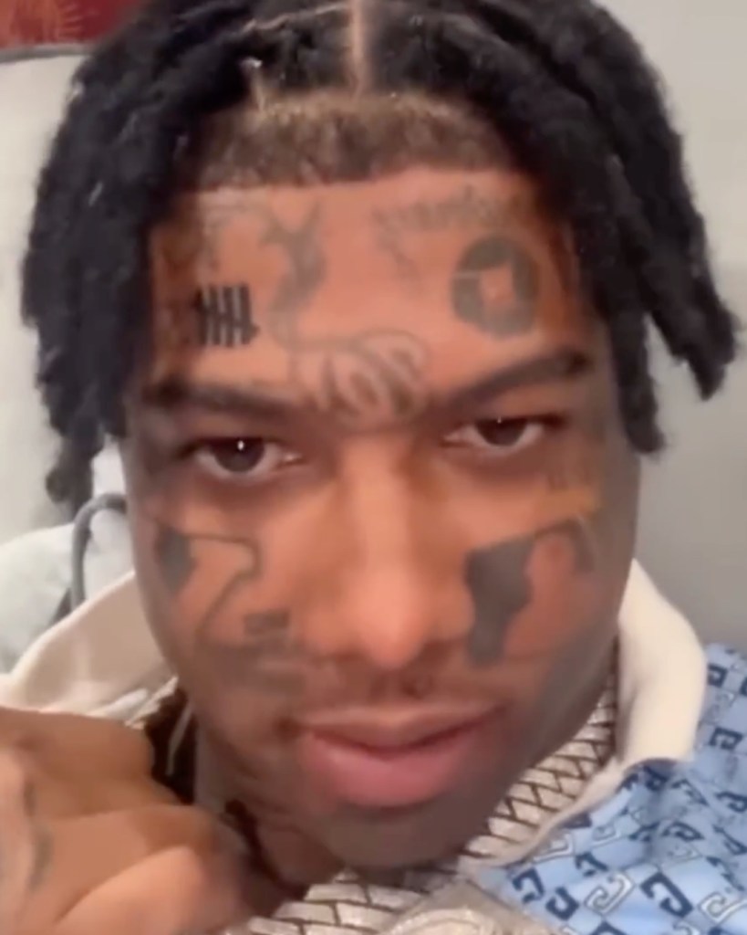 Close-up of Blueface with multiple facial tattoos, including brand logos, looking at the camera with braided hair.
