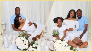 Singer 9ice poses in a white-themed maternity shoot with his pregnant partner and their daughter, surrounded by flowers and candles.