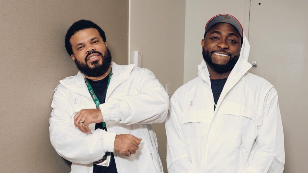 Davido and his manager Asa Asika pose side by side in matching white jackets, smiling against an indoor wall.