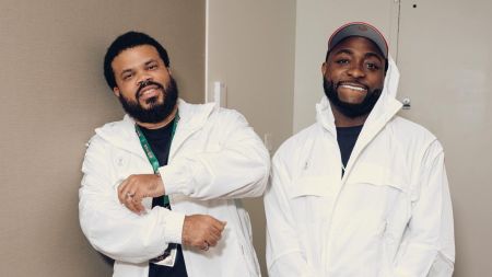 Davido and his manager Asa Asika pose side by side in matching white jackets, smiling against an indoor wall.