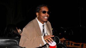 A$AP Rocky smiles while holding a red cup as he steps out of a car at night, wearing sunglasses, a beige suit and tie.