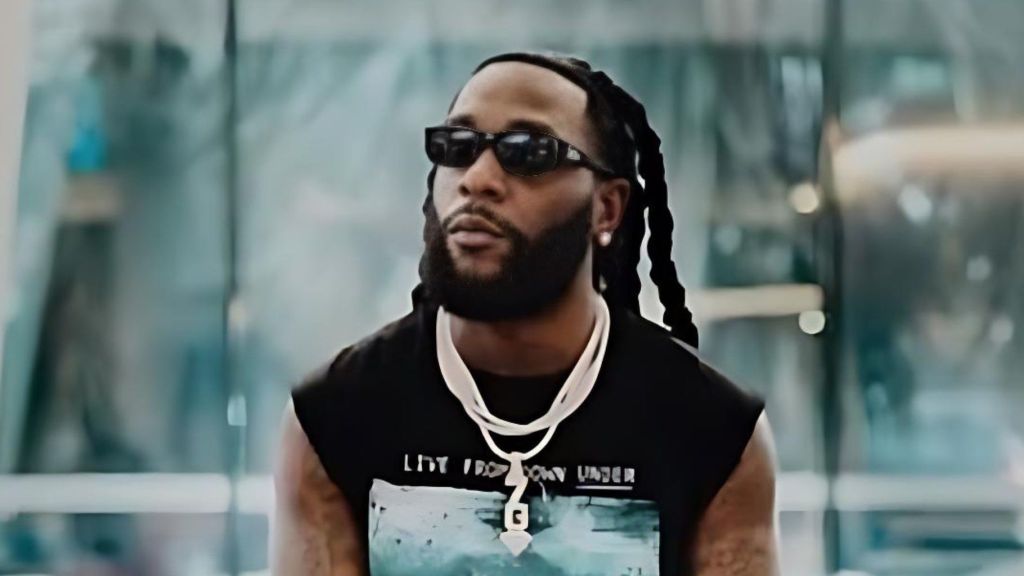 Burna Boy sits indoors wearing dark sunglasses, a black graphic sleeveless shirt and layered white necklaces, looking slightly upward with a calm expression.