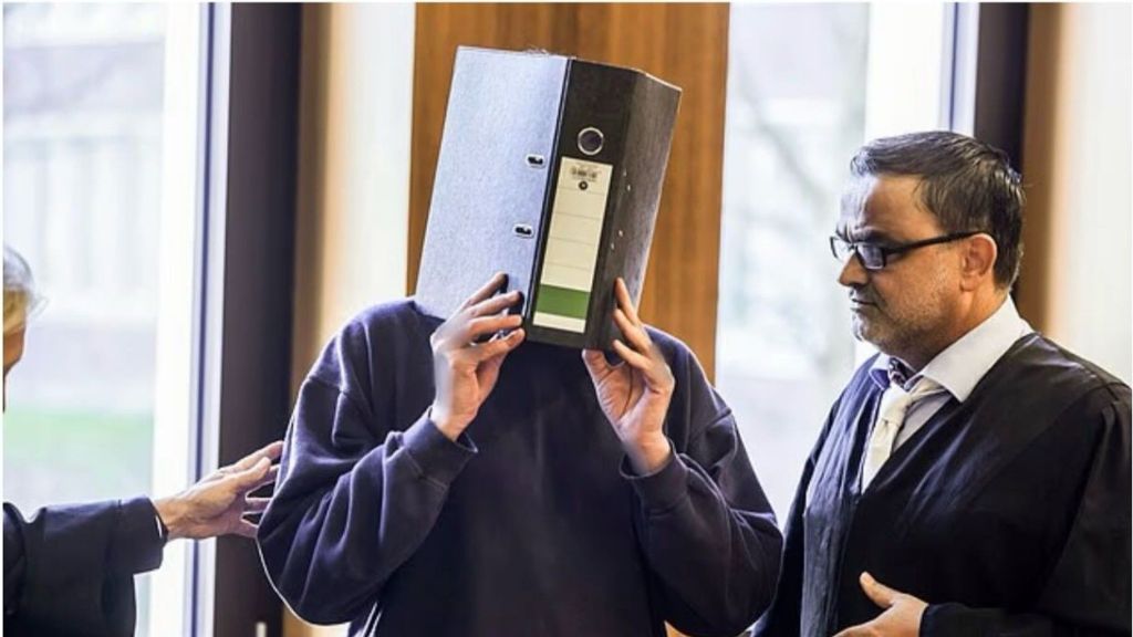 A man believed to be the convicted nurse stands in a courtroom, covering his face with a large black file folder while a lawyer in a black robe stands beside him.