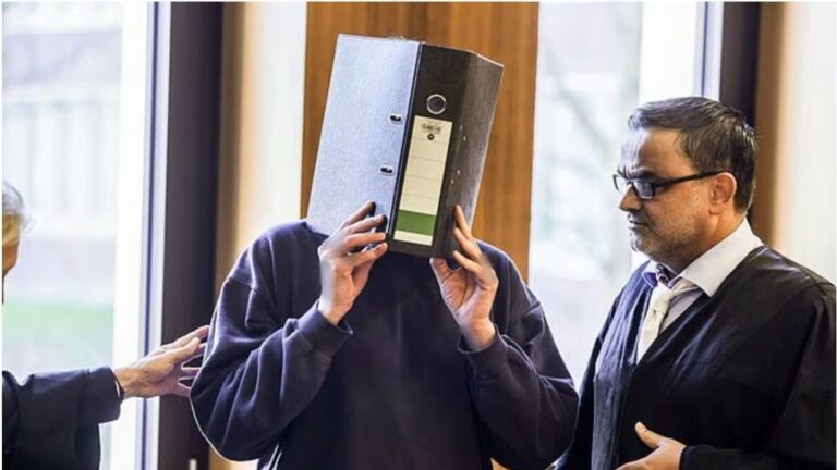 A man believed to be the convicted nurse stands in a courtroom, covering his face with a large black file folder while a lawyer in a black robe stands beside him.