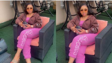 A young woman sits outdoors on a wicker chair, dressed in a brown hoodie and pink joggers, holding a phone. She wears tinted sunglasses, jewellery, and slippers, with a handbag placed beside the chair