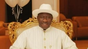 Goodluck Jonathan seated on an ornate gold-trimmed chair, wearing a white hat and white traditional outfit, smiling toward the camera.