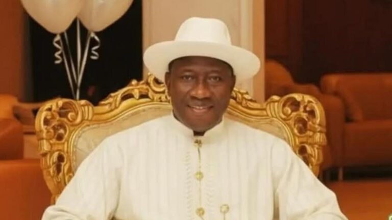 Goodluck Jonathan seated on an ornate gold-trimmed chair, wearing a white hat and white traditional outfit, smiling toward the camera.