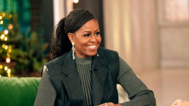 Michelle Obama smiling while seated on a talk show set, wearing long braided hair tied back, a grey sweater and dark vest, with soft lights and a blurred background behind her.
