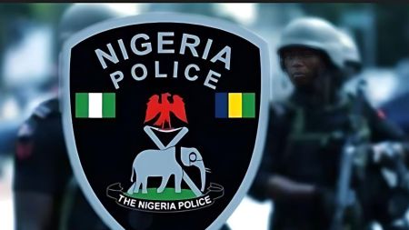 A close-up of the Nigeria Police Force emblem, featuring an eagle, crossed staves, and an elephant, with armed police officers blurred in the background.