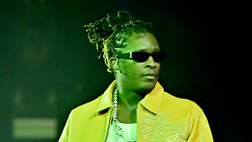 Young Thug performs on stage wearing a yellow jacket, dark sunglasses and diamond chain, with green stage lighting behind him.