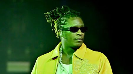 Young Thug performs on stage wearing a yellow jacket, dark sunglasses and diamond chain, with green stage lighting behind him.