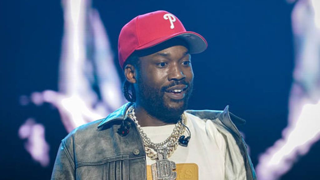 Meek Mill wearing a red cap and layered diamond chains while performing on stage.