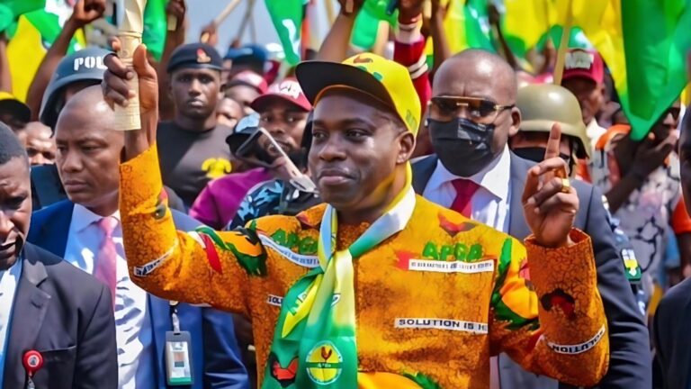 Chukwuma Soludo raises both hands while celebrating with supporters during a political rally, wearing a colourful APGA outfit and cap, surrounded by security and crowd holding party flags.