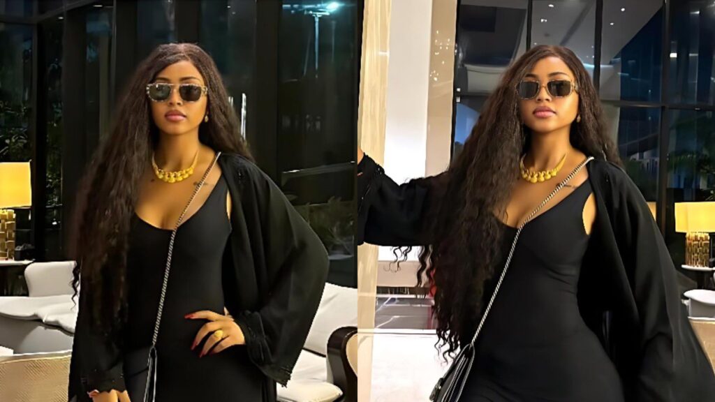 Regina Daniels poses indoors in a fitted black dress with a long black jacket, wearing sunglasses and a gold necklace, standing in a stylish modern lobby with warm lighting.