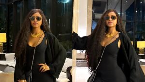 Regina Daniels poses indoors in a fitted black dress with a long black jacket, wearing sunglasses and a gold necklace, standing in a stylish modern lobby with warm lighting.