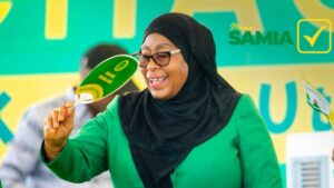 Tanzanian president Samia Suluhu Hassan smiles and waves a green CCM campaign fan while wearing a black hijab and green outfit at a rally, with the words “Mama Samia” in the background.