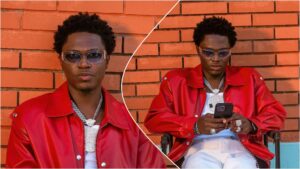 Spyro sits against an orange brick wall wearing a red leather jacket, white inner shirt, layered silver chains and rings, and clear-framed sunglasses; in one image he looks at the camera, and in the other he looks down at his phone.