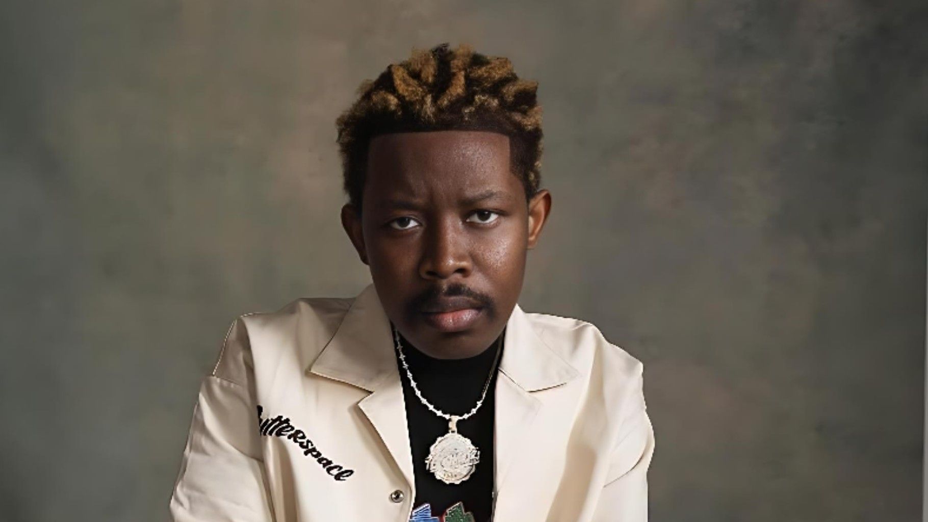 Nigerian music video director TG Omori poses for a studio portrait, wearing a cream jacket and silver chain, looking directly at the camera.