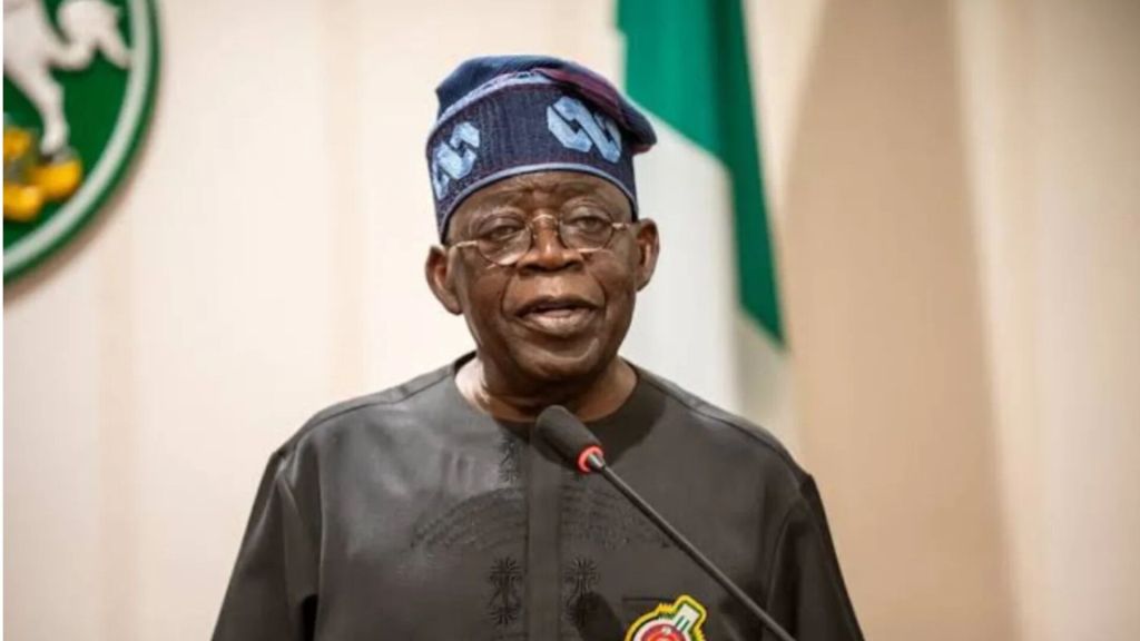 President Bola Ahmed Tinubu speaking at a podium while wearing a traditional cap and dark attire, with part of the Nigerian Coat of Arms visible in the background.