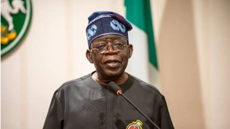 President Bola Ahmed Tinubu speaking at a podium while wearing a traditional cap and dark attire, with part of the Nigerian Coat of Arms visible in the background.